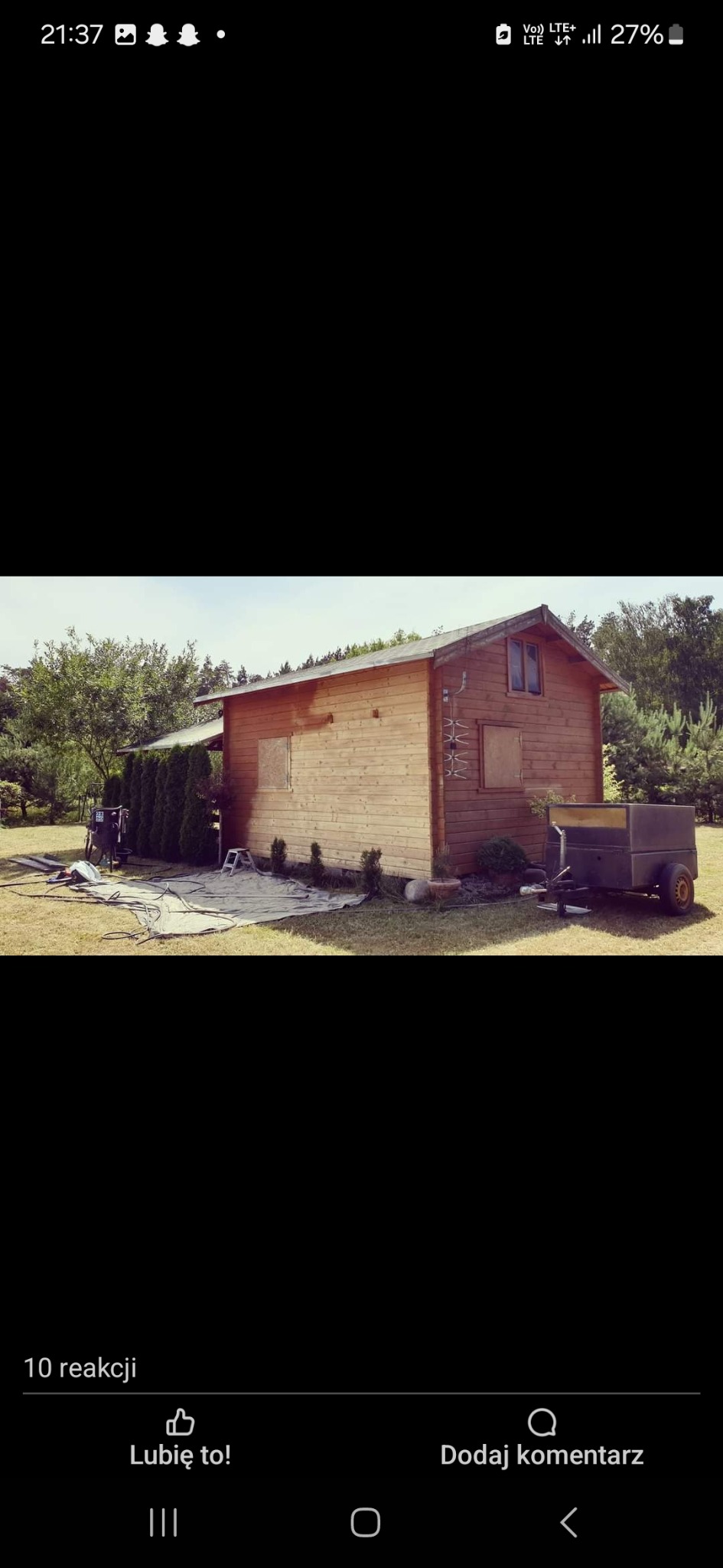 Drewniany domek letniskowy z przyczepką na trawniku, przygotowany do prac malarskich, z widoczną plandeką, narzędziami i drabinką.