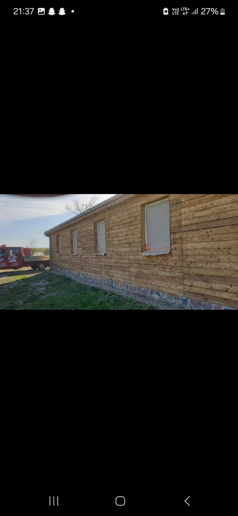 Drewniana elewacja budynku z zabezpieczonymi oknami przed malowaniem, widoczny fragment kamiennej podmurówki i trawnika, w tle czerwona laweta.