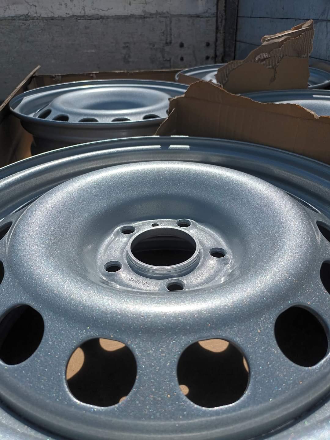 Świeżo pomalowane na srebrno felgi samochodowe w kartonowym opakowaniu, widoczne otwory montażowe i wentylacyjne.