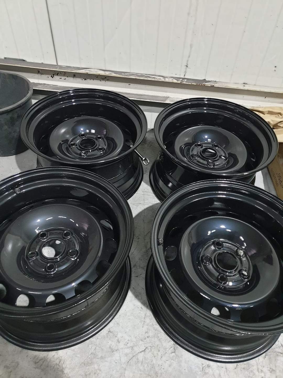 Cztery czarne, metalowe felgi samochodowe po renowacji z Kruszwicy, ustawione na szarej podłodze w warsztacie.