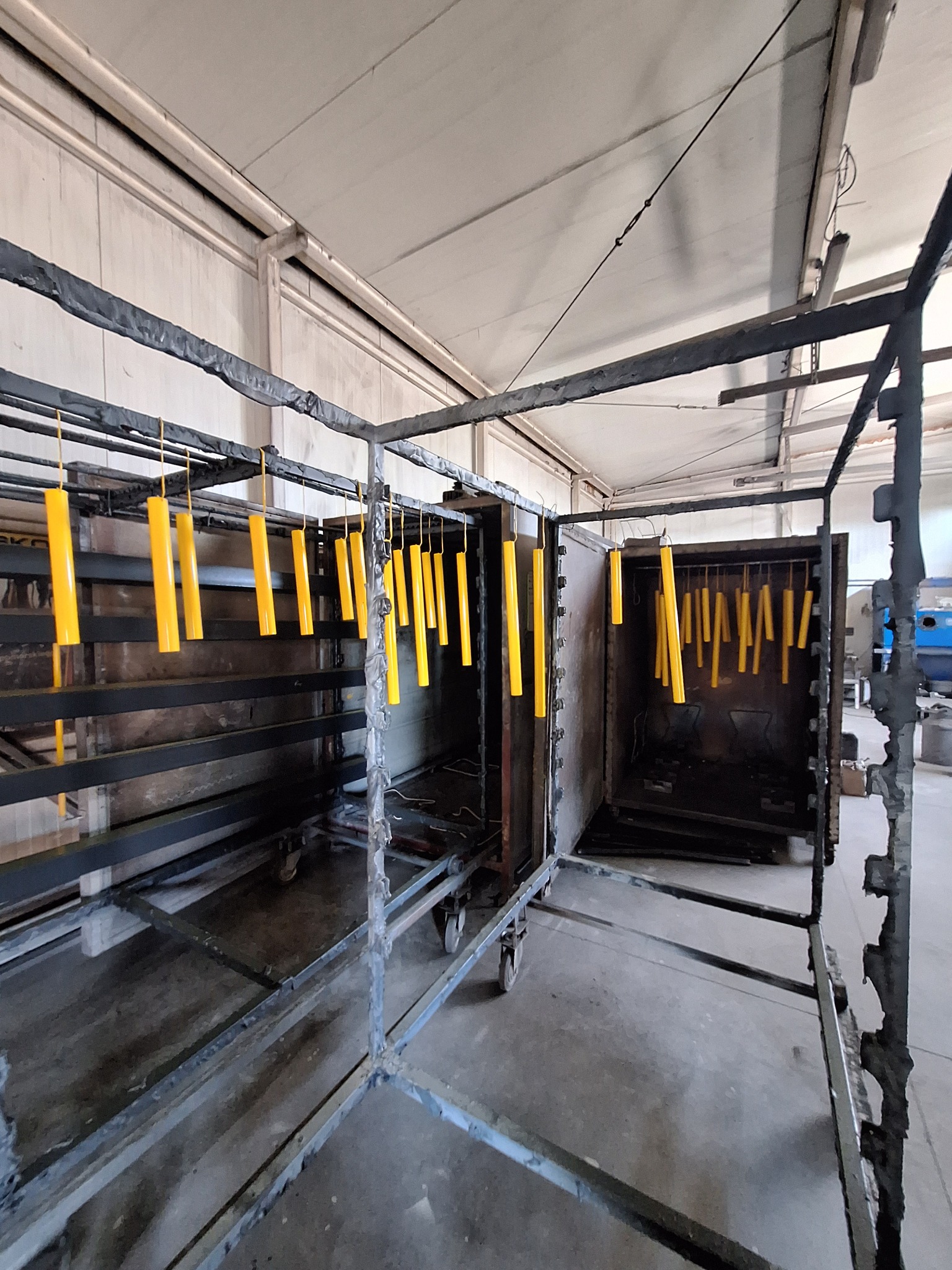 Żółte, cylindryczne elementy zawieszone na hakach wewnątrz metalowej konstrukcji, prawdopodobnie przygotowane do procesu nakładania powłoki; widoczny fragment pieca.