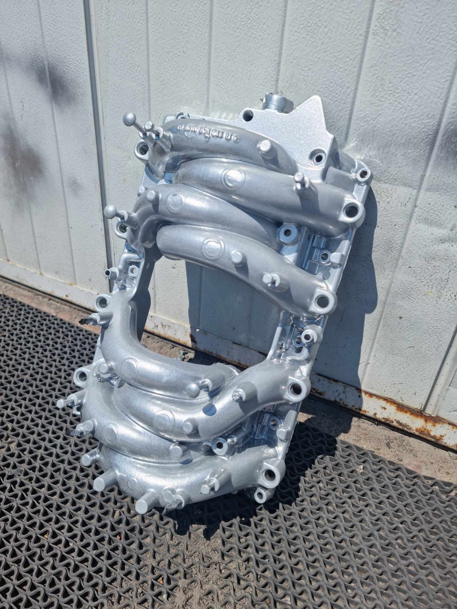 Kolektor ssący pomalowany proszkowo na srebrno, leżący na gumowej macie, widoczne detale i struktura powłoki lakierniczej.