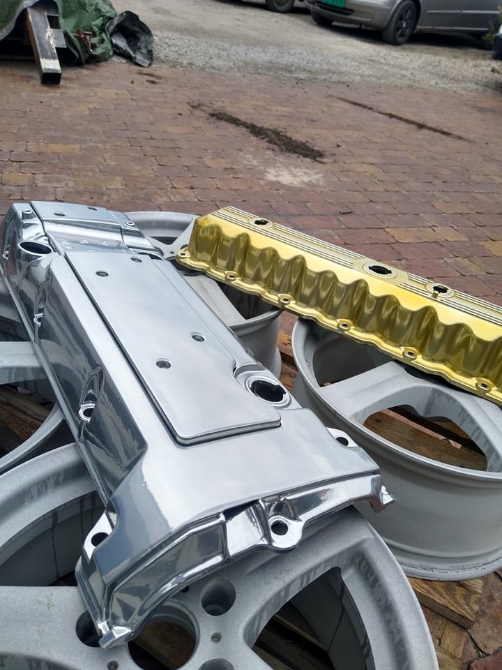 Dwie pokrywy zaworów samochodowych, jedna srebrna, druga złota, leżące na szarych felgach samochodowych na tle brukowanej powierzchni.