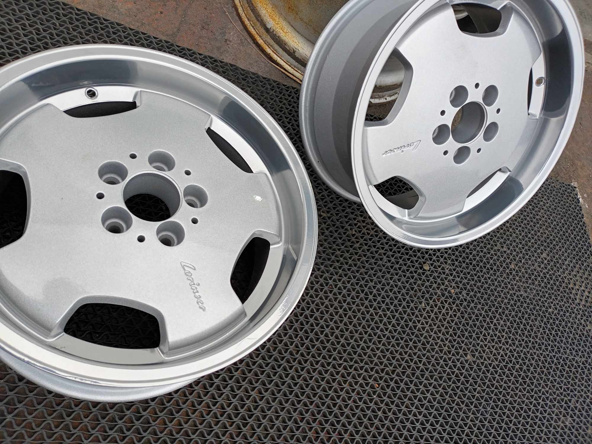 Dwa srebrne felgi aluminiowe z wygrawerowanym napisem 'Lorinser' leżące na gumowej macie, z widocznym fragmentem zardzewiałej felgi w tle.