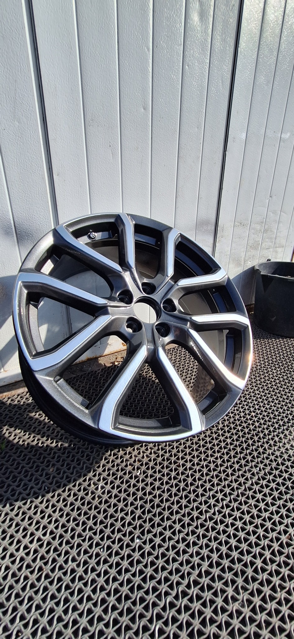 Felga aluminiowa samochodowa, pomalowana proszkowo na kolor grafitowy z srebrnymi akcentami, leży na gumowej wycieraczce na tle szarej ściany z paneli.