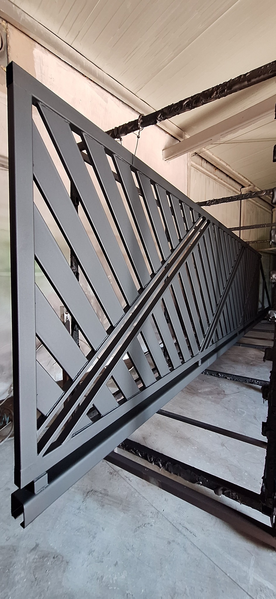 Metalowa brama z ukośnymi prętami, świeżo polakierowana na szaro, zawieszona pod sufitem w warsztacie, widoczne ślady po lakierowaniu proszkowym.