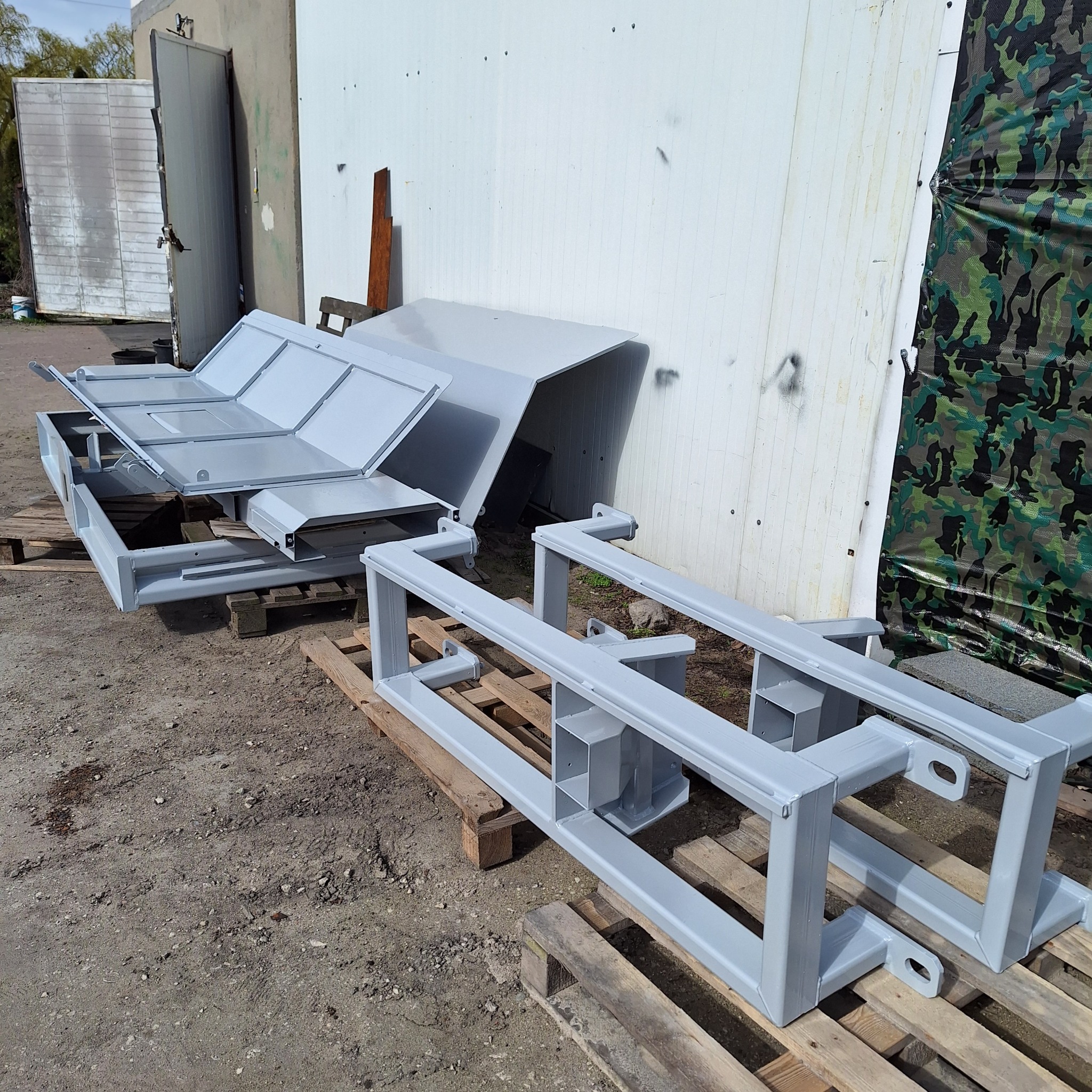 Elementy konstrukcyjne, prawdopodobnie do przyczepy lub maszyny rolniczej, świeżo po malowaniu proszkowym, leżące na paletach przed budynkiem z widocznymi śladami zabrudzeń.