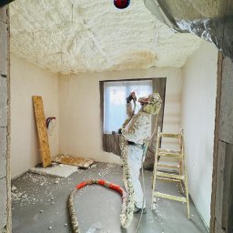 RAF - Development Rafał Kościuk - Pracownik w kombinezonie ochronnym aplikuje pianę izolacyjną na sufit wewnątrz budynku. Widoczne narzędzia, drabina i fragment okna.