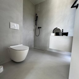 RAF - Development Rafał Kościuk - Minimalistyczna łazienka z szarymi ścianami i podłogą, wisząca toaleta, czarna bateria prysznicowa i umywalka na białej szafce, odpływ liniowy w podłodze.