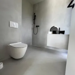 Minimalistyczna łazienka z szarymi ścianami i podłogą, wisząca toaleta, czarna bateria prysznicowa i umywalka na białej szafce, odpływ liniowy w podłodze.