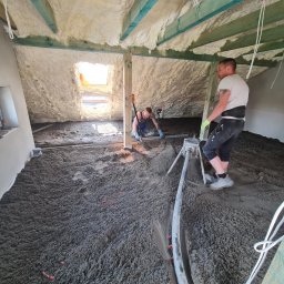 RAF - Development Rafał Kościuk - Prace w trakcie w Obornikach Śląskich: w pomieszczeniu na poddaszu, po ociepleniu pianką, dwóch mężczyzn rozprowadza wylewkę cementową, używając poziomicy i listwy wibracyjnej.