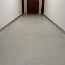 RAF - Development Rafał Kościuk - Wnętrze korytarza z wykończoną podłogą w odcieniach szarości, białe ściany z szarymi listwami przypodłogowymi, brązowe drzwi z elementami dekoracyjnymi i gaśnica na ścianie.