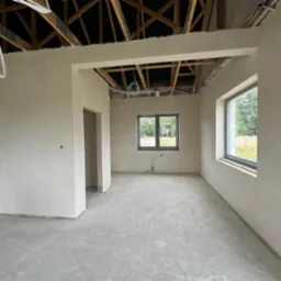 Surowe wnętrze domu w trakcie prac wykończeniowych. Ściany pokryte świeżym tynkiem, widoczne okna z szarymi ramami, surowy beton na podłodze i odsłonięta drewniana konstrukcja dachu z widocznymi...