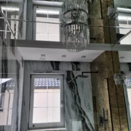 Elegancka łazienka z marmurowymi płytami imitującymi żyły, lustrzanym sufitem odbijającym kryształowy żyrandol i złotą mozaiką na ścianie kabiny prysznicowej, widok na okno z zewnątrz.