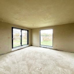 RAF - Development Rafał Kościuk - Wnętrze po tynkowaniu w Obornikach Śląskich: ściany pokryte świeżym tynkiem, widoczne okna i instalacje elektryczne. Podłoga surowa, przygotowana do dalszych prac.