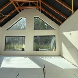 RAF - Development Rafał Kościuk - Wnętrze domu w trakcie prac wykończeniowych: ściany po tynkowaniu, widoczne okna o nietypowym kształcie, podłoga przygotowana pod wylewkę. Elementy drewniane dachu.