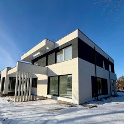 RAF - Development Rafał Kościuk - Nowoczesny dom z elewacją w bieli i czerni, widoczny w zimowej scenerii. Minimalistyczny design, duże okna, płaski dach. Częściowo ośnieżony teren wokół budynku.