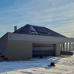 RAF - Development Rafał Kościuk - Nowoczesny dom z garażem w zimowej scenerii, widok z zewnątrz. Szara elewacja, panele słoneczne na dachu, błękitne niebo z promieniami słońca.