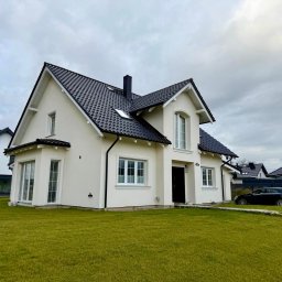 RAF - Development Rafał Kościuk - Biały dom jednorodzinny z czarnym dachem i białymi oknami, otoczony zielonym trawnikiem. Widoczne elementy wykończenia elewacji i detale architektoniczne. Pochmurne niebo w tle.