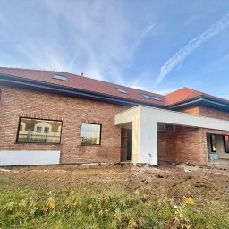 RAF - Development Rafał Kościuk - Nowoczesny dom z czerwonej cegły w trakcie budowy, z widocznymi oknami i fragmentem białego zadaszenia. Ujęcie z poziomu gruntu, błękitne niebo w tle.