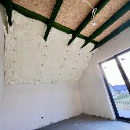 RAF - Development Rafał Kościuk - Poddasze z widoczną izolacją pianką PUR między krokwiami, surowy stan ścian, okno dachowe z widokiem na zewnątrz. Detal: widoczne mocowania pianki.