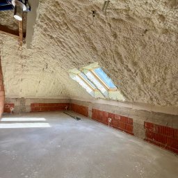 RAF - Development Rafał Kościuk - Poddasze po ociepleniu pianką PUR, widoczne okna dachowe i instalacje. Ściany z cegły częściowo ocieplone. Szara, betonowa podłoga. Oświetlenie naturalne.