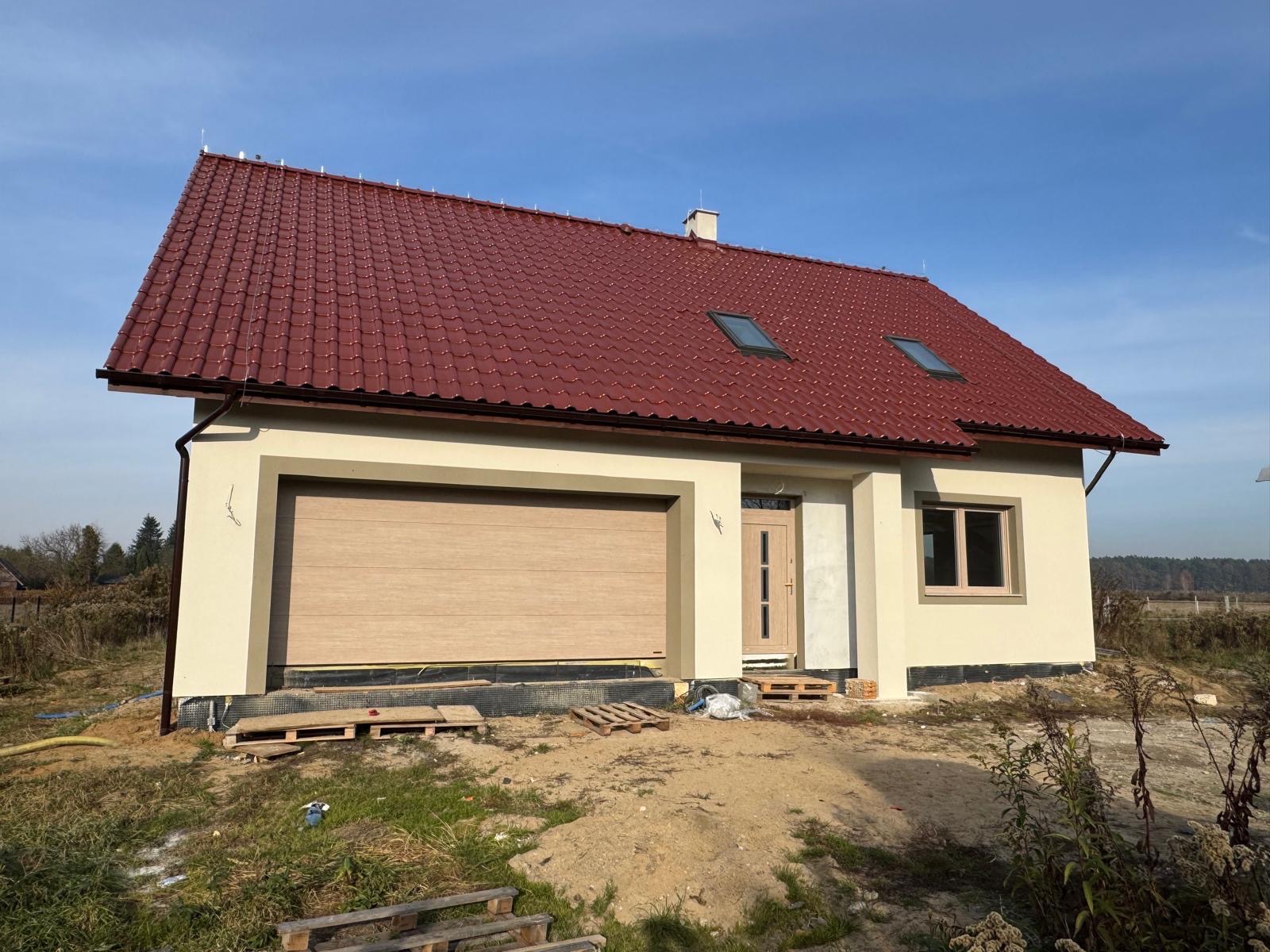 Nowo wybudowany dom jednorodzinny z beżową elewacją, brązową bramą garażową i czerwoną dachówką, widoczne palety i folia zabezpieczająca fundamenty.