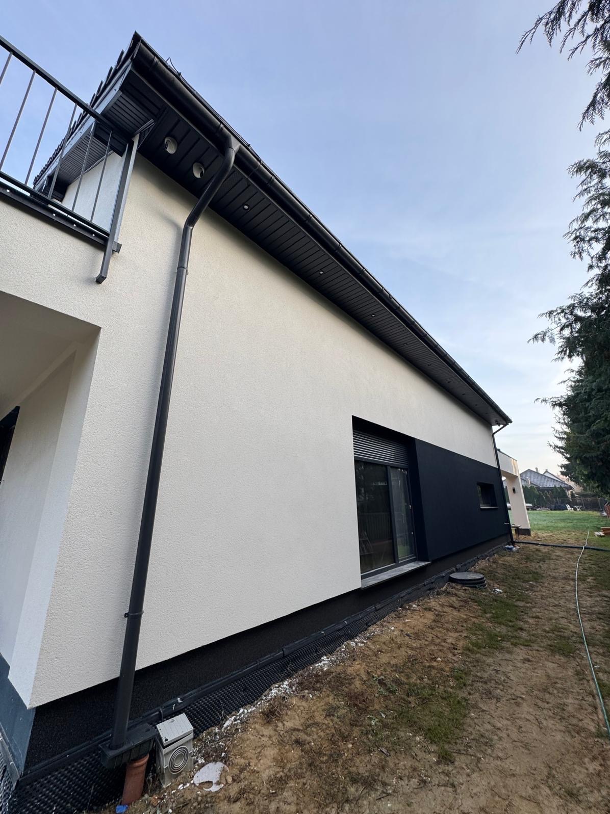 Nowoczesna elewacja domu jednorodzinnego z fragmentem balkonu, widoczna biała i grafitowa kolorystyka, orynnowanie oraz drenaż przy fundamentach.