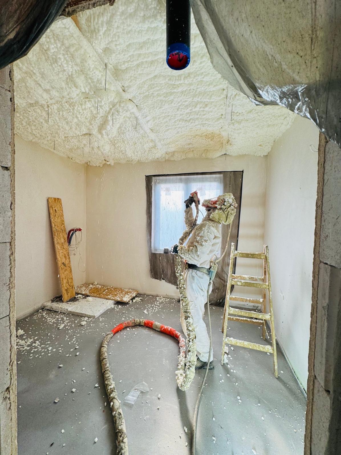 Pracownik w kombinezonie ochronnym aplikuje pianę izolacyjną na sufit wewnątrz budynku. Widoczne narzędzia, drabina i fragment okna.