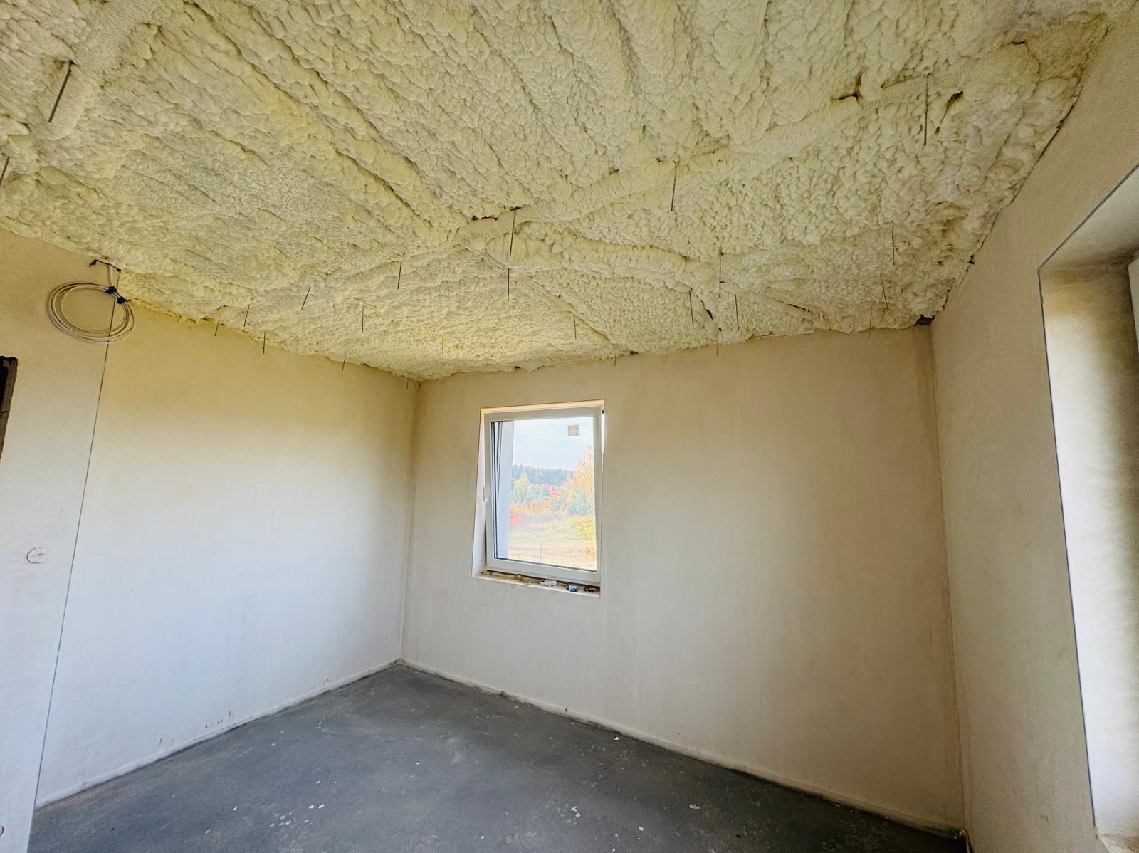 Wnętrze pokoju z surowymi ścianami i sufitem pokrytym grubą warstwą pianki izolacyjnej, z widocznymi metalowymi mocowaniami, okno z widokiem na jesienne drzewa.