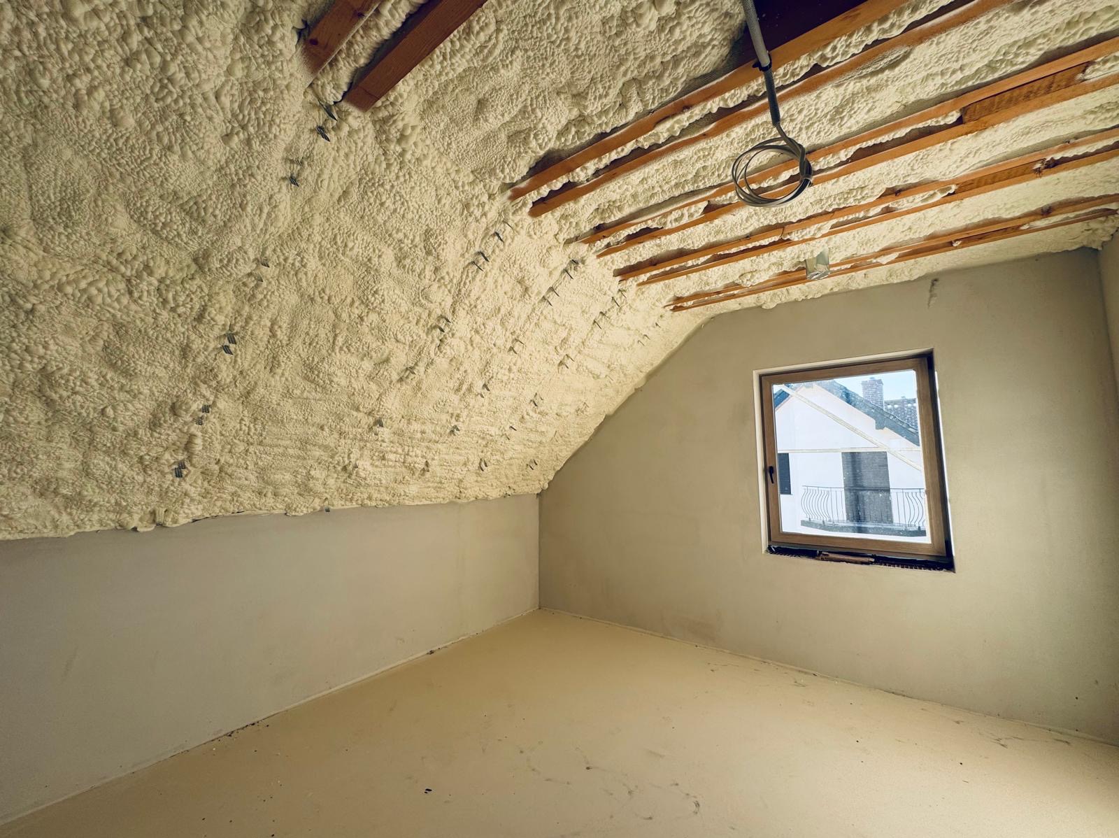 Poddasze po ociepleniu pianką poliuretanową, widoczne drewniane belki stropowe i elementy montażowe, ściany pomalowane na szaro, okno z widokiem na budynek z balkonem.