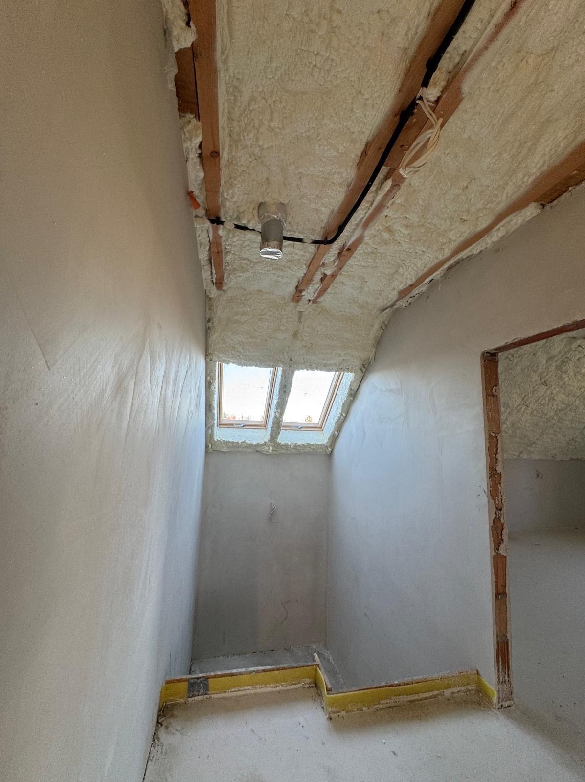 Poddasze w trakcie prac wykończeniowych: ściany pokryte gładzią, sufit ocieplony pianą PUR z widocznymi belkami drewnianymi, dwa okna dachowe, żółta taśma zabezpieczająca na podłodze.