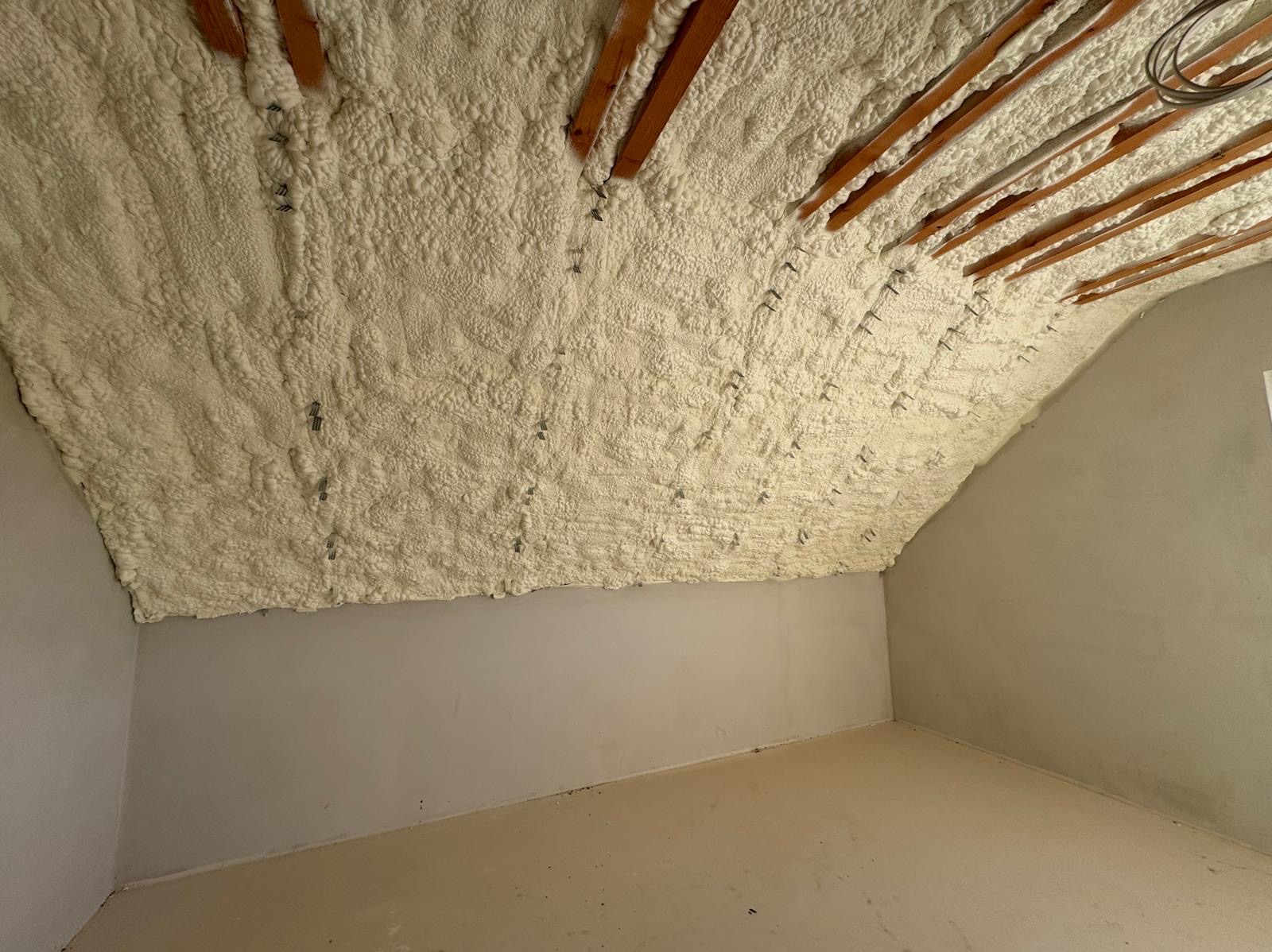 Poddasze ocieplone pianą PUR, widoczne drewniane belki stropowe i metalowe zaczepy, surowe ściany w odcieniach szarości, podłoga w jasnym beżu.