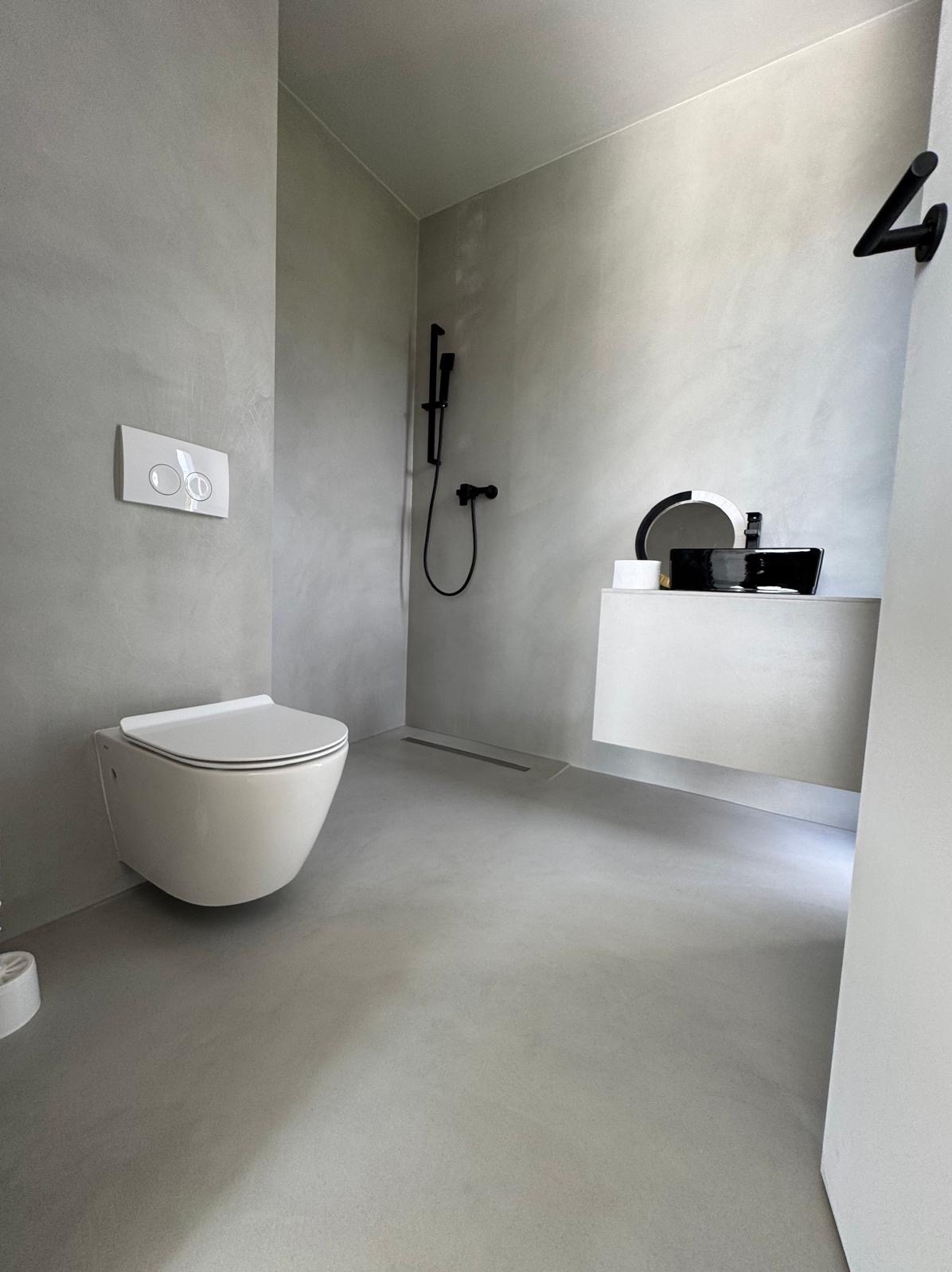 Minimalistyczna łazienka z szarymi ścianami i podłogą, wisząca toaleta, czarna bateria prysznicowa i umywalka na białej szafce, odpływ liniowy w podłodze.