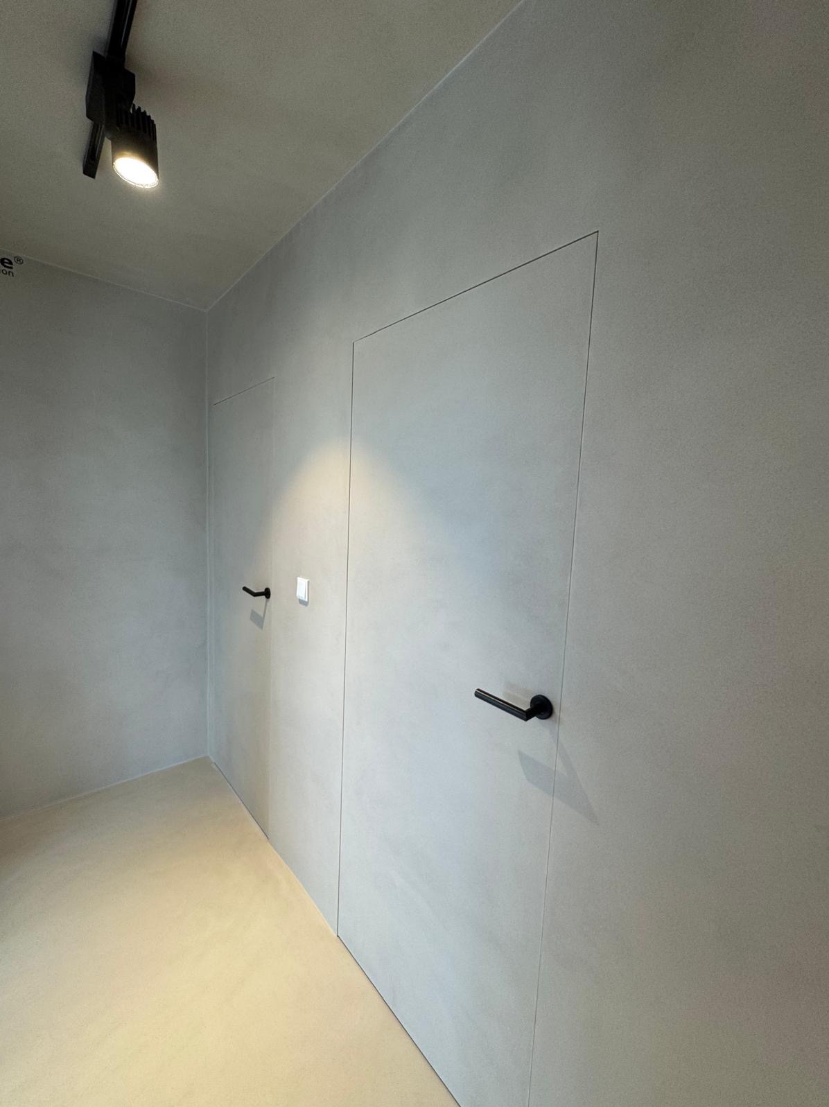 Minimalistyczne wnętrze z gładkimi, szarymi ścianami, ukrytymi drzwiami z czarnymi klamkami i punktowym oświetleniem sufitowym, tworzące jednolitą, nowoczesną powierzchnię.