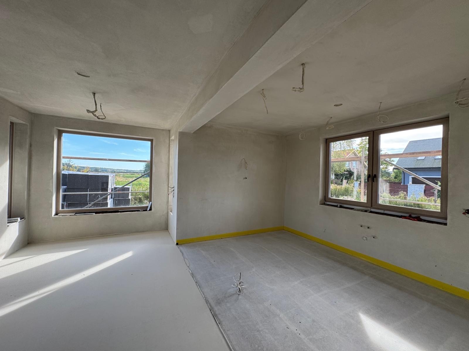 Wnętrze surowego domu w stanie deweloperskim, ściany i sufit pokryte świeżym, szarym tynkiem, na podłodze wylewka, okna z widokiem na zewnątrz, zabezpieczone żółtą taśmą przypodłogową.