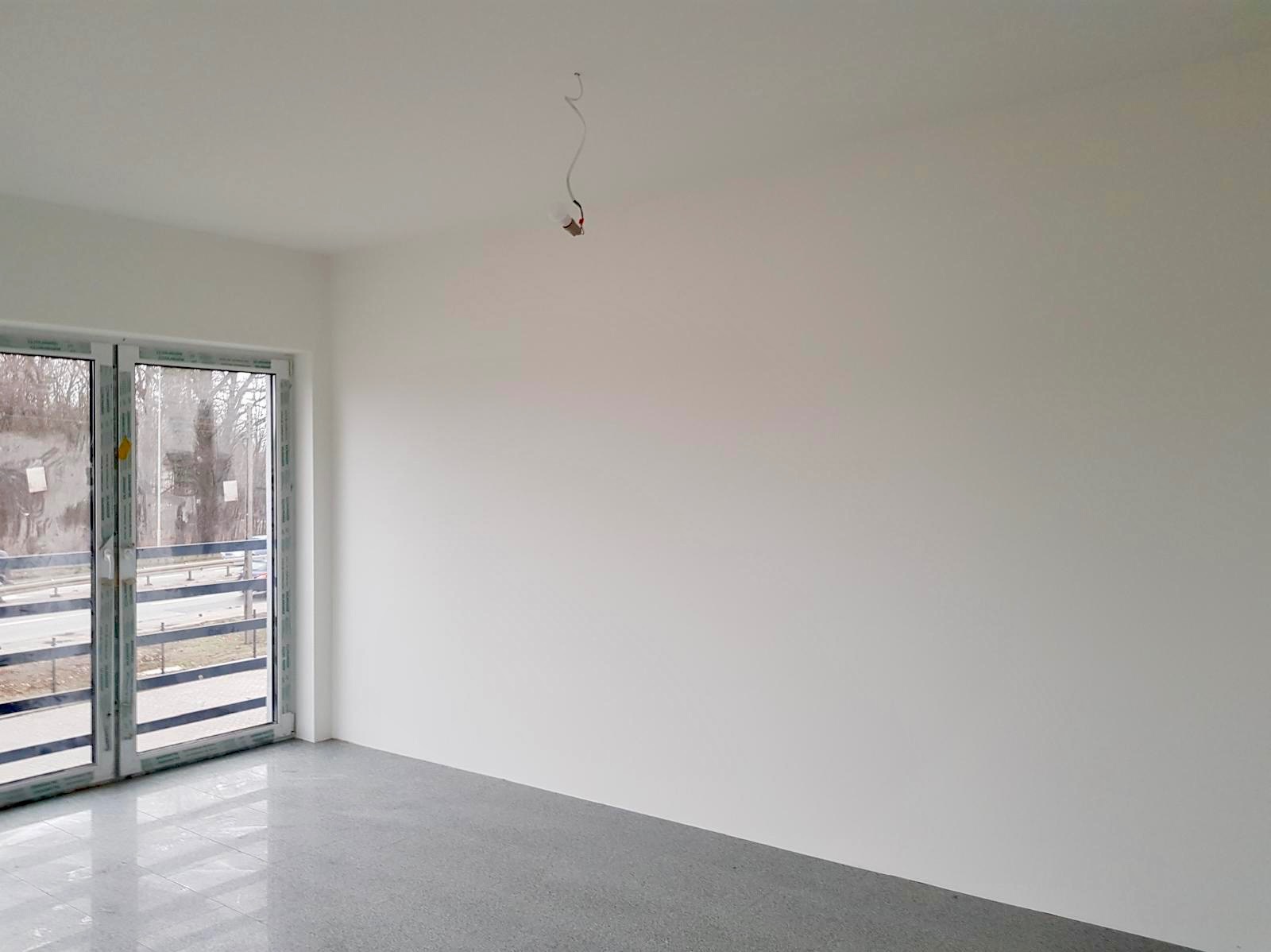Wnętrze pokoju po pracach wykończeniowych; gładkie, białe ściany, szara podłoga, okno balkonowe z widokiem na ulicę, wystający kabel ze ściany.