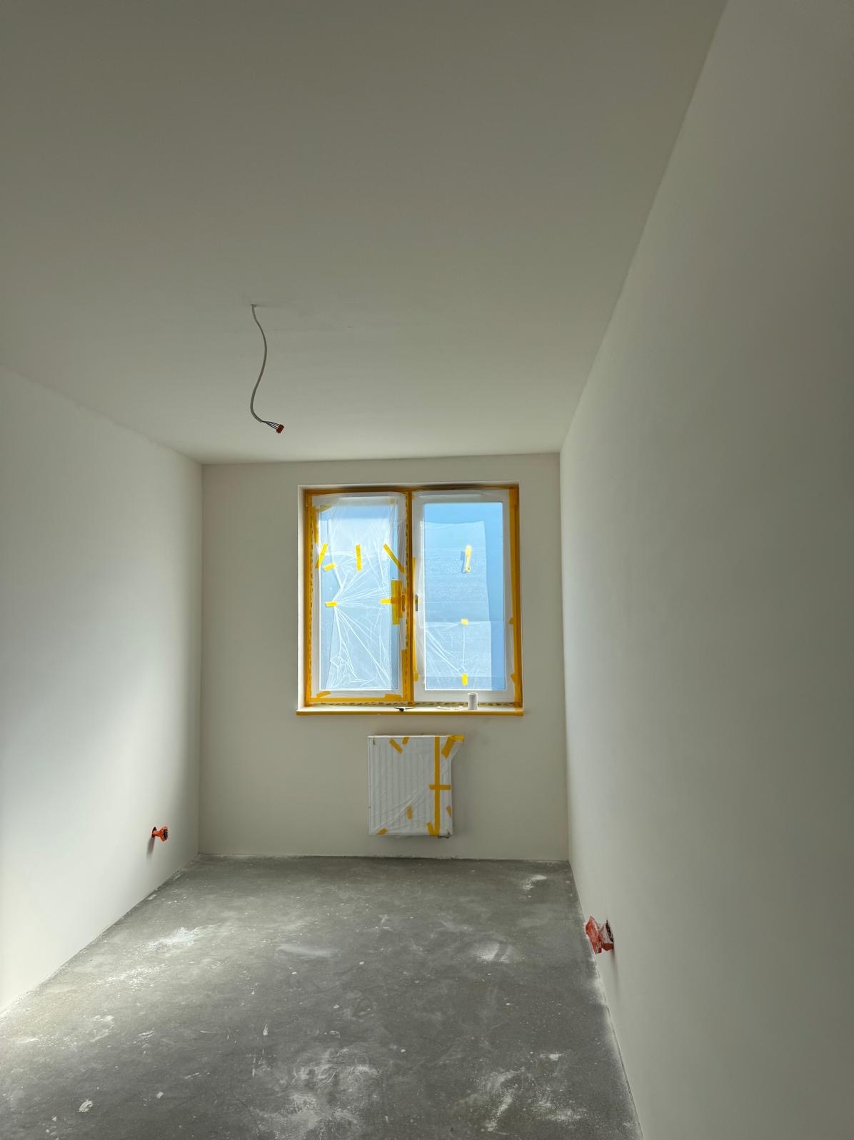 Pokój w stanie deweloperskim po wylewce, z oknem i grzejnikiem zabezpieczonym folią i taśmą malarską, przygotowany do prac wykończeniowych ścian i sufitu. Widoczne wystające kable elektryczne.