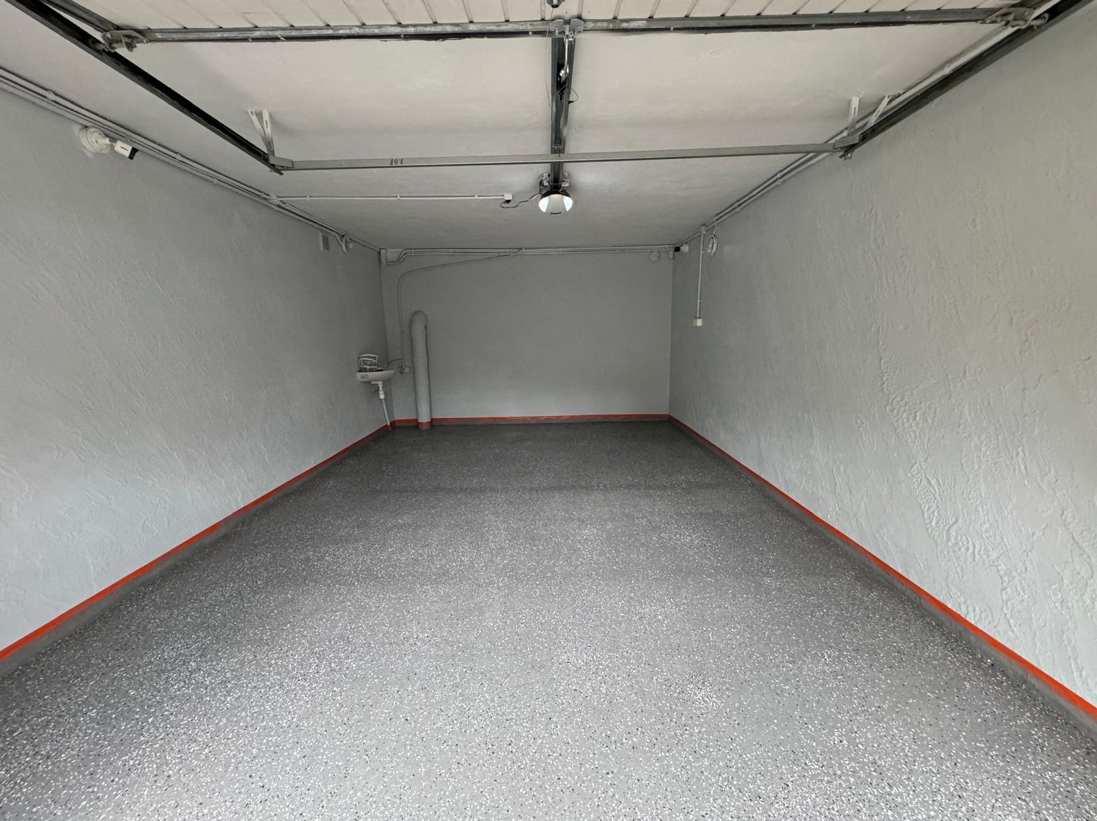 Wnętrze garażu po remoncie: szare ściany o wyraźnej fakturze, szara posadzka z brokatem, pomalowane na pomarańczowo listwy przypodłogowe, widoczna instalacja elektryczna i umywalka.