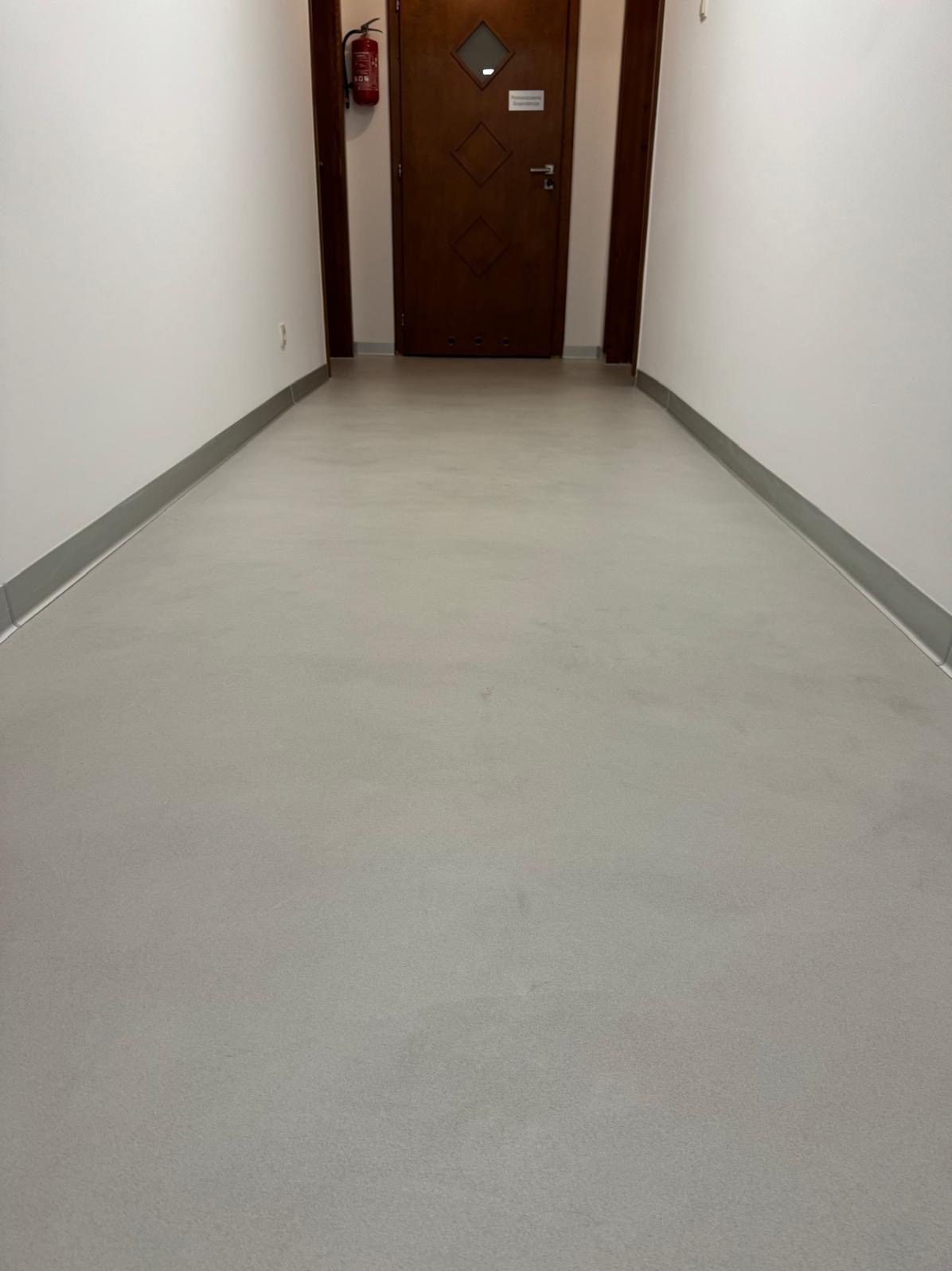 Wnętrze korytarza z wykończoną podłogą w odcieniach szarości, białe ściany z szarymi listwami przypodłogowymi, brązowe drzwi z elementami dekoracyjnymi i gaśnica na ścianie.