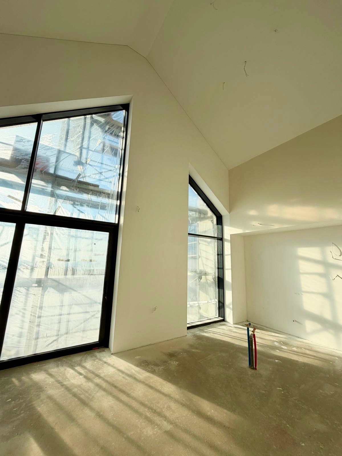 Wnętrze z gładzią na ścianach i suficie, duże okna o nietypowym kształcie, przygotowane do dalszych prac wykończeniowych, widoczne instalacje na podłodze.