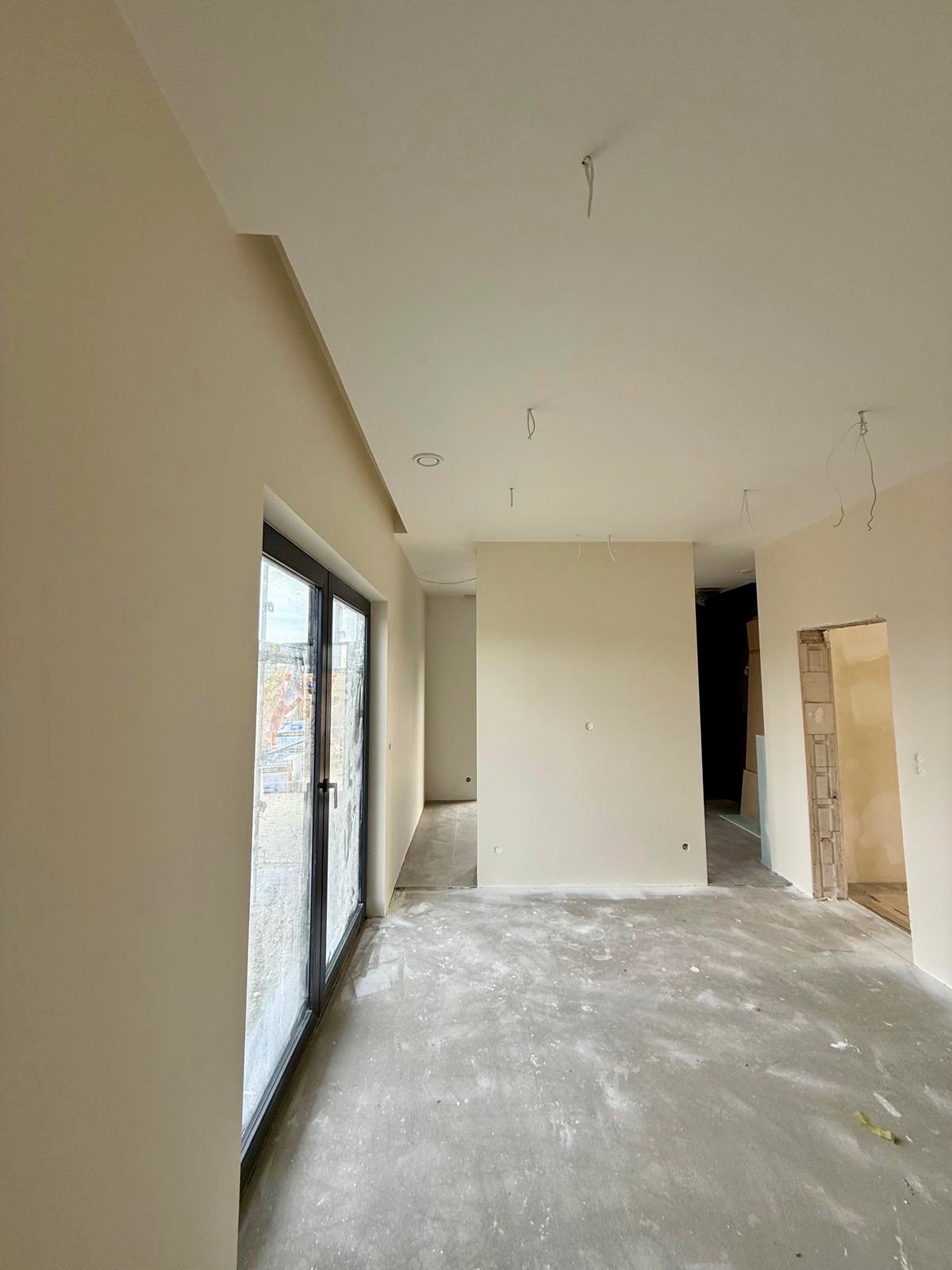 Wnętrze po gładziowaniu ścian i sufitu, z widocznymi przewodami elektrycznymi. Duże okno z czarną ramą i surowa podłoga. Ściany w odcieniach beżu.