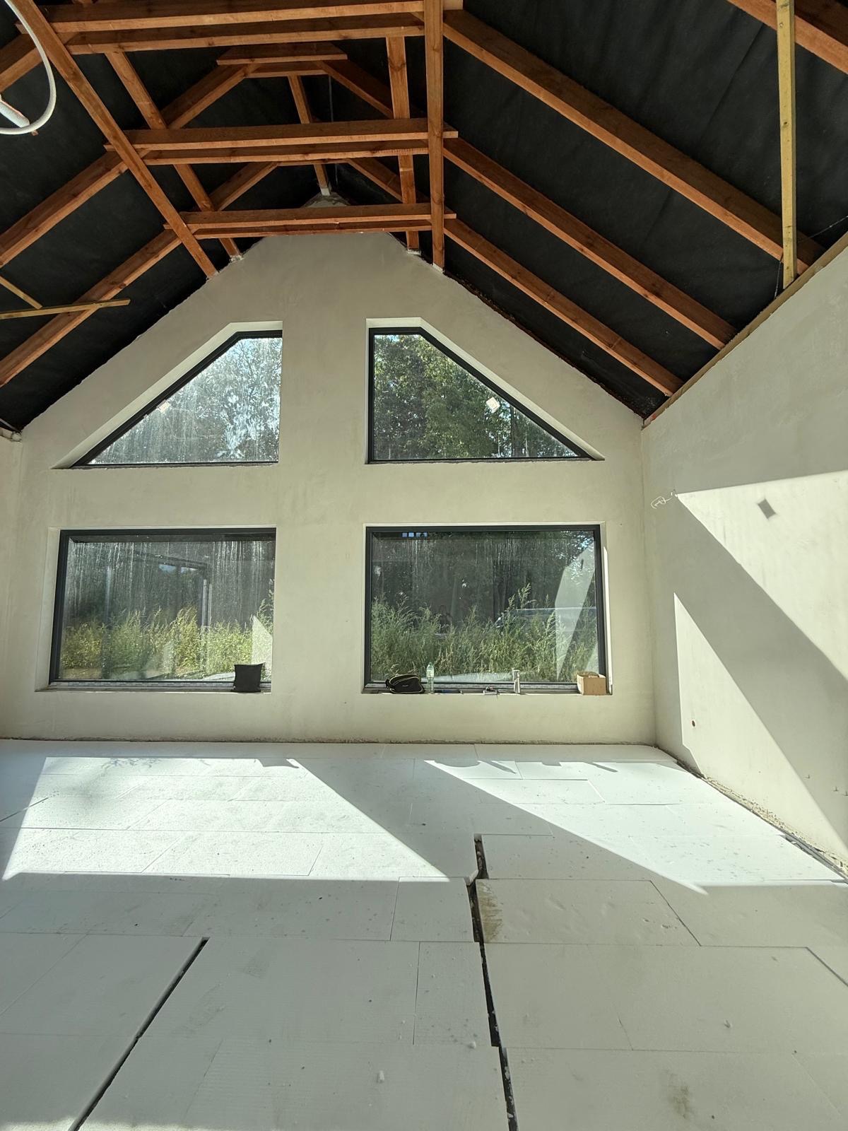 Wnętrze domu w trakcie prac wykończeniowych: ściany po tynkowaniu, widoczne okna o nietypowym kształcie, podłoga przygotowana pod wylewkę. Elementy drewniane dachu.