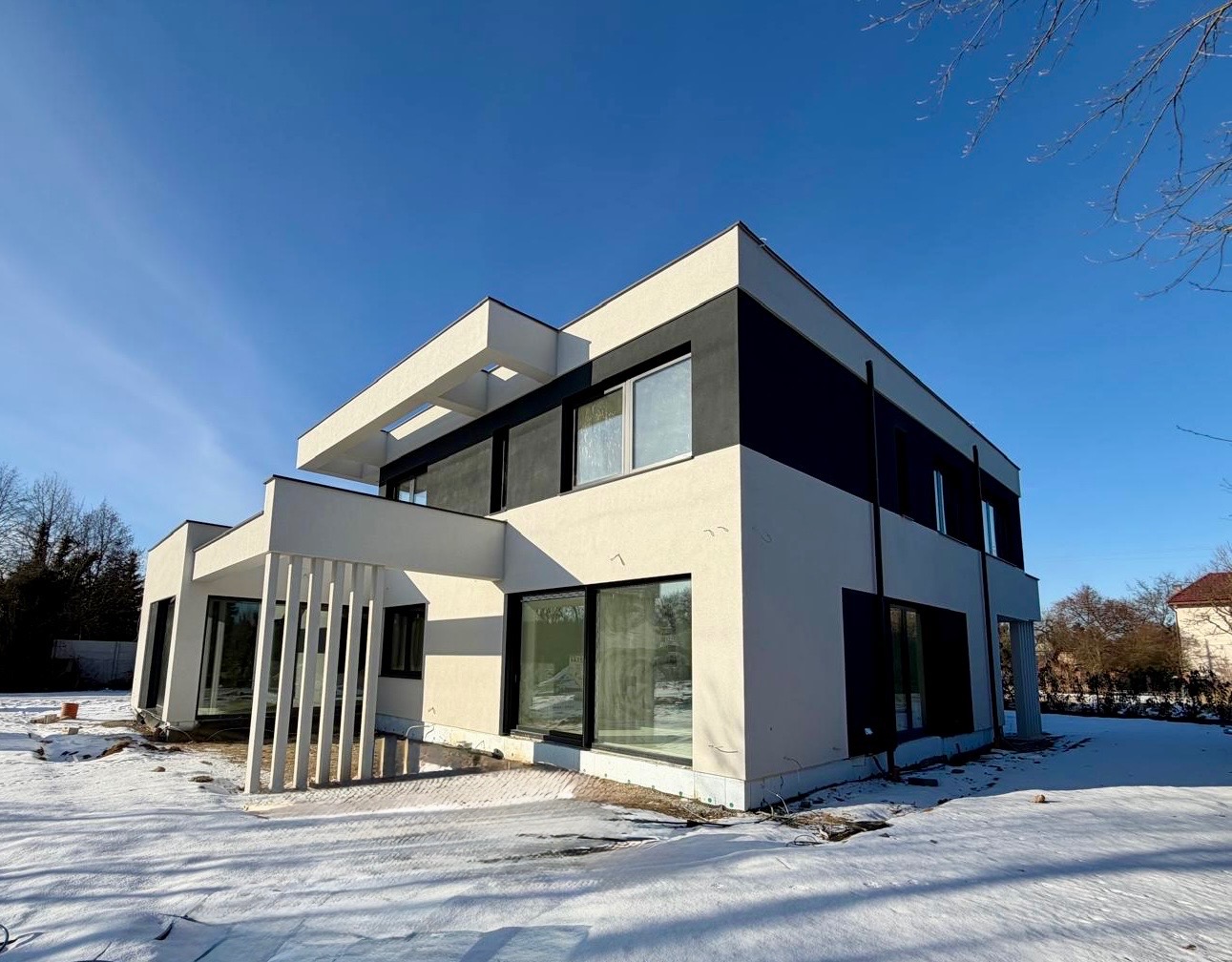 Nowoczesny dom z elewacją w bieli i czerni, widoczny w zimowej scenerii. Minimalistyczny design, duże okna, płaski dach. Częściowo ośnieżony teren wokół budynku.