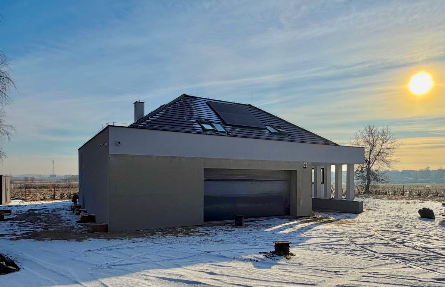 Nowoczesny dom z garażem w zimowej scenerii, widok z zewnątrz. Szara elewacja, panele słoneczne na dachu, błękitne niebo z promieniami słońca.