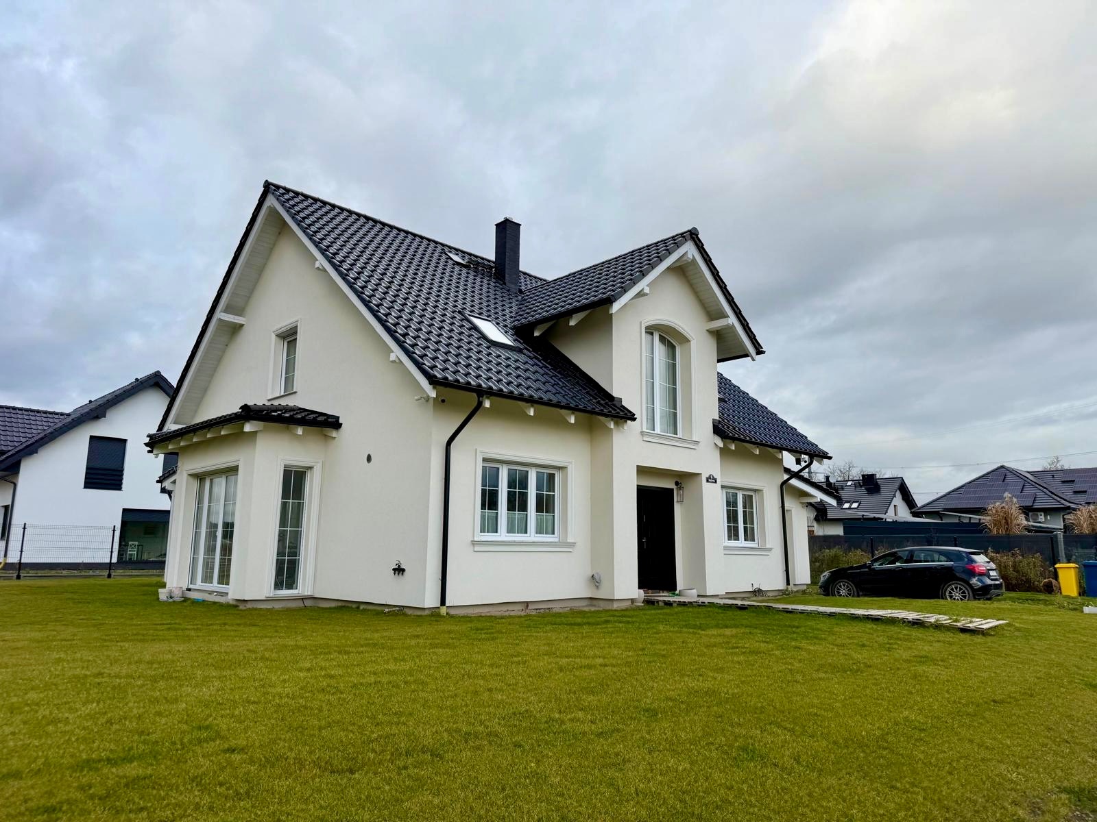 Biały dom jednorodzinny z czarnym dachem i białymi oknami, otoczony zielonym trawnikiem. Widoczne elementy wykończenia elewacji i detale architektoniczne. Pochmurne niebo w tle.