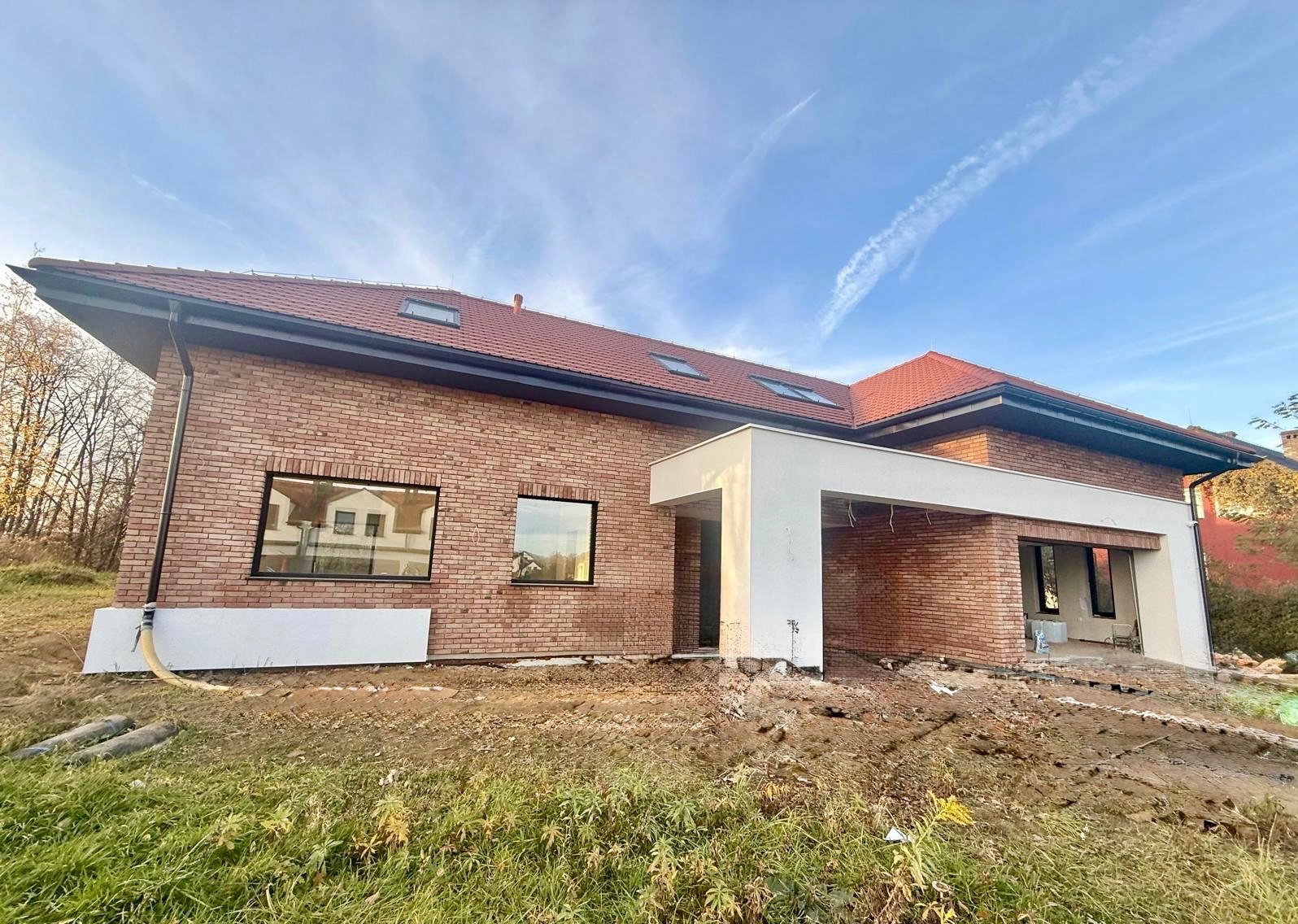 Nowoczesny dom z czerwonej cegły w trakcie budowy, z widocznymi oknami i fragmentem białego zadaszenia. Ujęcie z poziomu gruntu, błękitne niebo w tle.