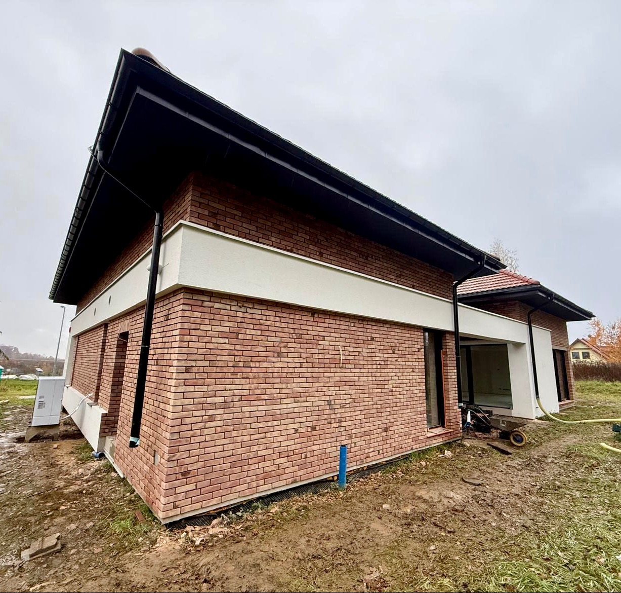 Nowoczesny dom z ceglaną elewacją, czarnym dachem i białym pasem dekoracyjnym. Widoczne rynny i fragment otoczenia. Ujęcie z boku, pochmurne niebo.