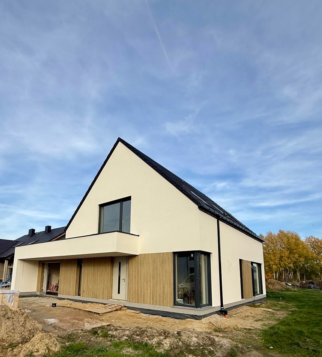 Nowoczesny dom jednorodzinny z jasną elewacją i drewnianym panelem na dole, czarne okna i dach, błękitne niebo w tle. Widok z zewnątrz, w trakcie wykańczania.