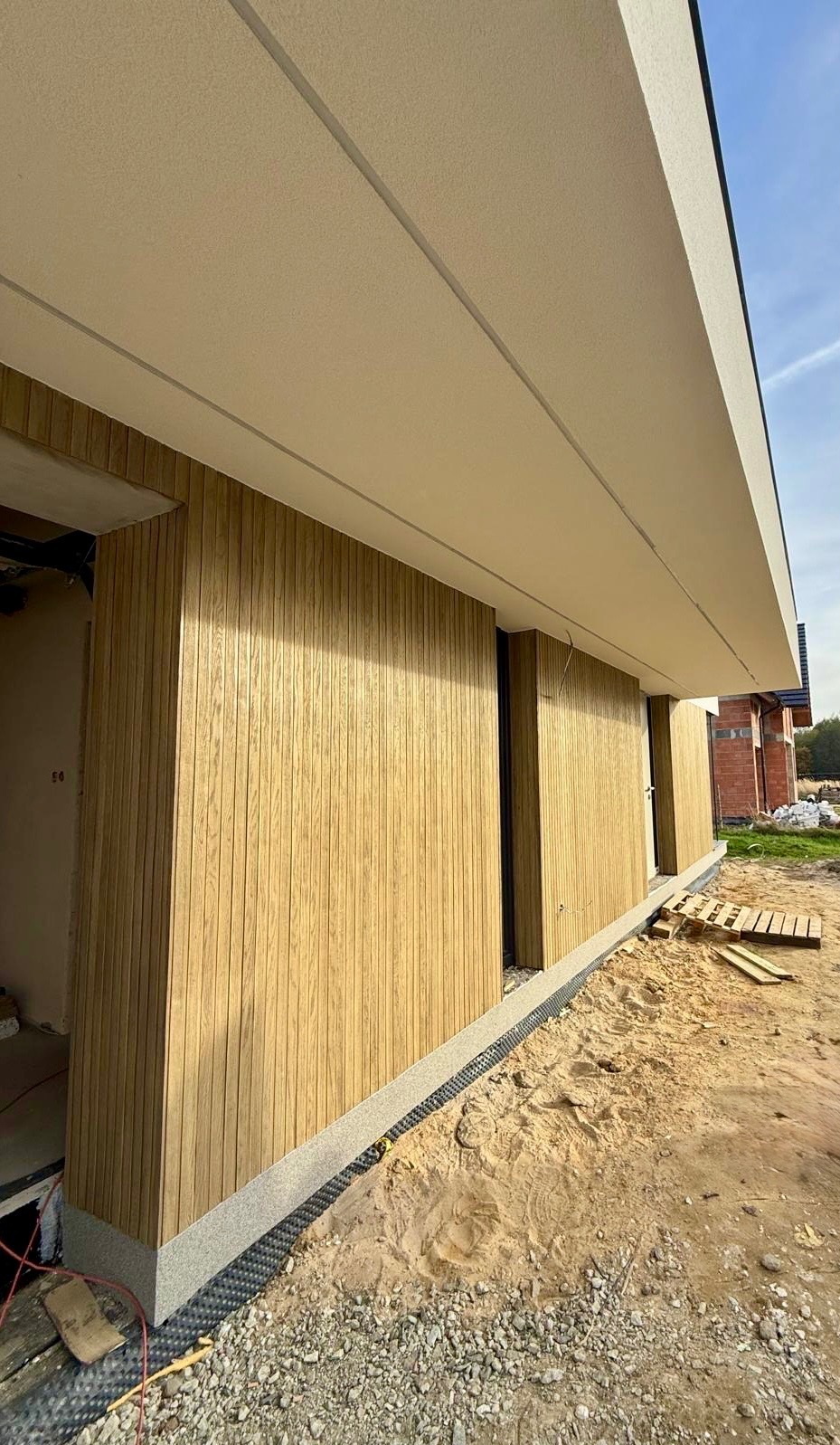 Nowoczesna elewacja domu jednorodzinnego z drewnianą okładziną i jasnym, gładkim podbitką. Widoczne detale wykończenia i teren wokół budynku w trakcie prac.